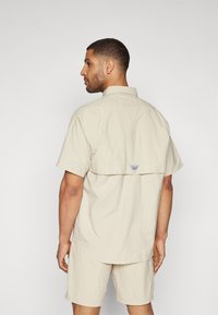 Camicia a maniche corte di un leggero beige con orlo curvato, caratterizzata da un piccolo tag blu sul retro. Vestibilità semplice e rilassata con texture morbida.