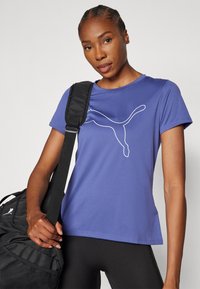 T-shirt atlético roxa com um logotipo de gato branco, mangas curtas e um corte relaxado. Mochila de mão preta com uma superfície texturizada e alça.