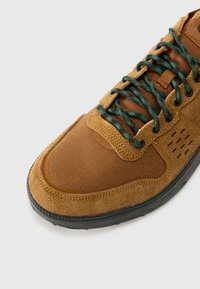 Brune semsket og mesh sneakers med grønne snørebånd, rund tå, teksturerte paneler og en svart gummisåle med detaljer.
