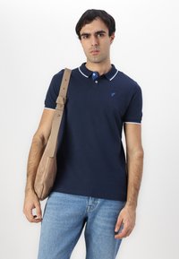 Navy polo shirt with white trim, short sleeves, and a small blue logo. Model wears light blue jeans and carries a beige shoulder bag.