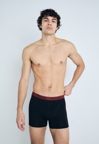 Črne bokserice z rdečim elastičnim pasom, na katerem je logotip "BJÖRN BORG". Imajo gladko teksturo in oprijet kroj. Model je brez majice.