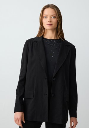Black jacket made of lightweight fabric, featuring a notched collar, two front pockets, and two black buttons. Worn over a black top with embellishments.