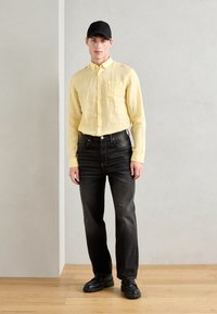 Yellow linen button-up shirt with chest pocket, paired with black jeans and black chunky shoes. Model wears a black cap. Neutral background.