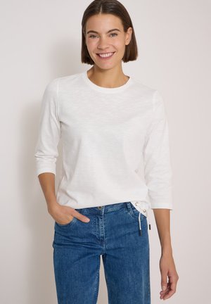Femme souriante aux cheveux courts et bruns, portant un haut blanc à manches longues et un jean bleu, une main dans la poche, debout devant un fond uni.