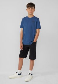 Blauwe t-shirt met korte mouwen en een rond logo, gecombineerd met knielange zwarte shorts en witte sneakers met marineblauwe accenten.