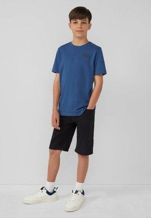 Blauwe t-shirt met korte mouwen en een rond logo, gecombineerd met knielange zwarte shorts en witte sneakers met marineblauwe accenten.