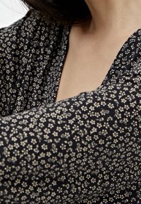 Czarna bluzka z tkaniny z drobnymi białymi kwiatowymi wzorami, głębokim dekoltem w kształcie litery V i miękką, zwiewną teksturą. Zbliżenie na obszar klatki piersiowej.