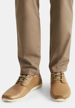 Hnědé kožené boty s hladkým povrchem, kulatou špičkou, světlými tkaničkami a jemným bočním prošitím. Spárovány s béžovými kalhotami.