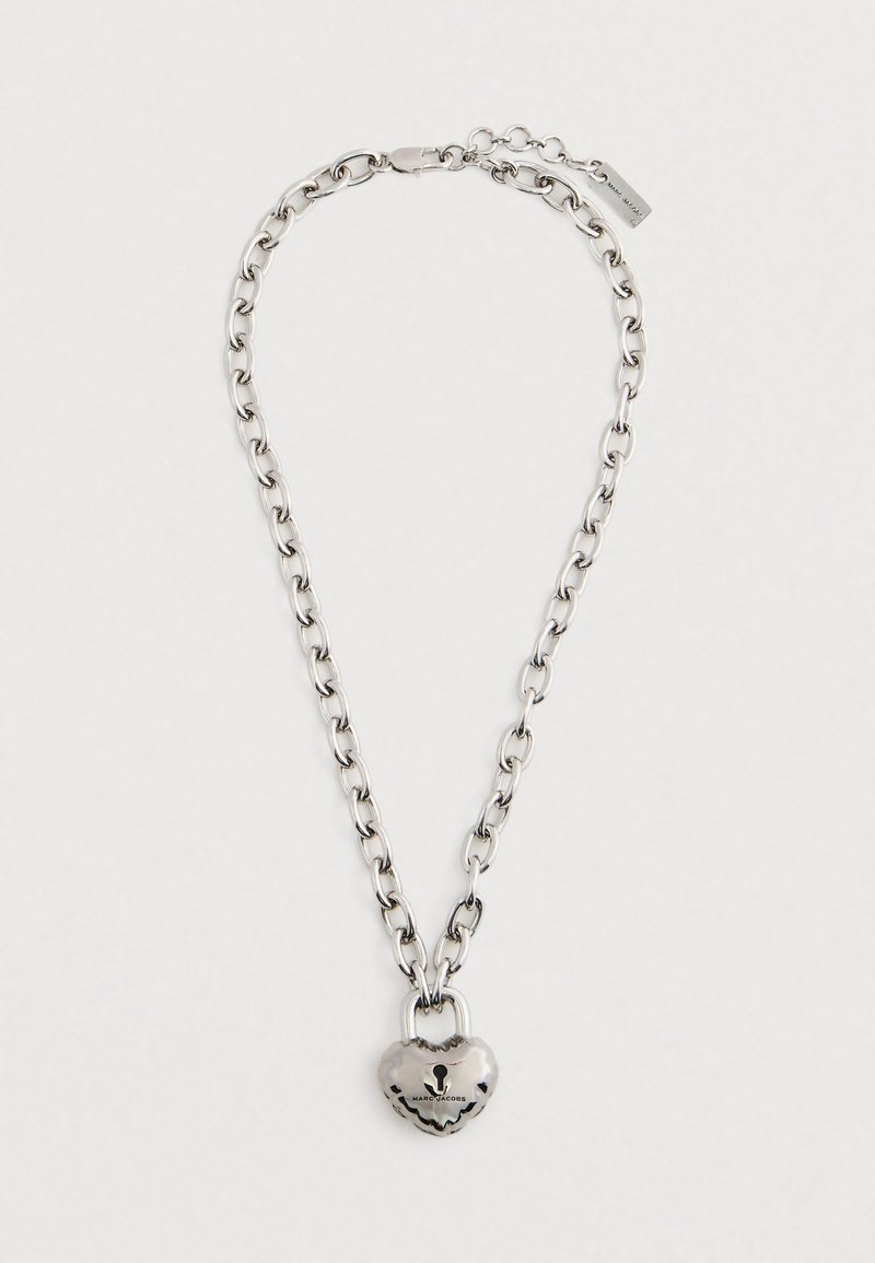 Srebrna verižica z obeskom v obliki srca, ki ima gladko teksturo in graviran logotip. Oblikovana za nastavljivo velikost.