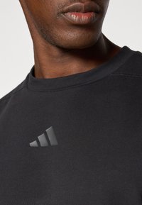 T-shirt noir à manches courtes en coton, avec un logo Adidas en gris texturé sur la partie gauche de la poitrine.