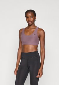 Sportowy stanik w mauve'owym kolorze, o gładkiej fakturze, z szerokimi ramiączkami i czarnym logo. W komplecie z czarnymi legginsami o wysokim stanie.