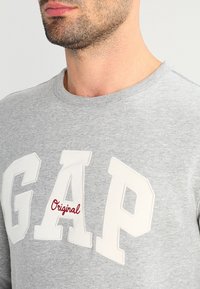 Hombre vistiendo un sudadera de cuello redondo en gris claro con grandes letras blancas "GAP" y el texto rojo "Original" bordado en el pecho.