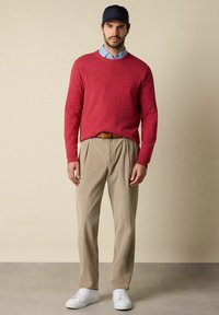 Roter Strickpullover, hellblaues Hemd mit Kragen darunter, beigefarbene Cordhose, weiße Sneakers, marineblaues Cap. Slim Fit, minimalistisches Design.
