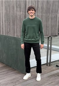 Zelený pletený svetr s texturovaným horním panelem, černé rovné kalhoty a bílé tenisky, stojící na dřevěné palubě.