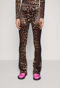 Legíny s leopardím potiskem z lesklého materiálu s rozšířeným střihem, v odstínech hnědé a zlaté, doplněné o růžové sandály na šedém pozadí.