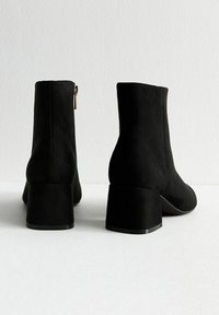 Zwarte suède enkelboots met een blokhak, voorzien van een zijrits en een gladde textuur, van achteren getoond met een minimalistisch ontwerp.