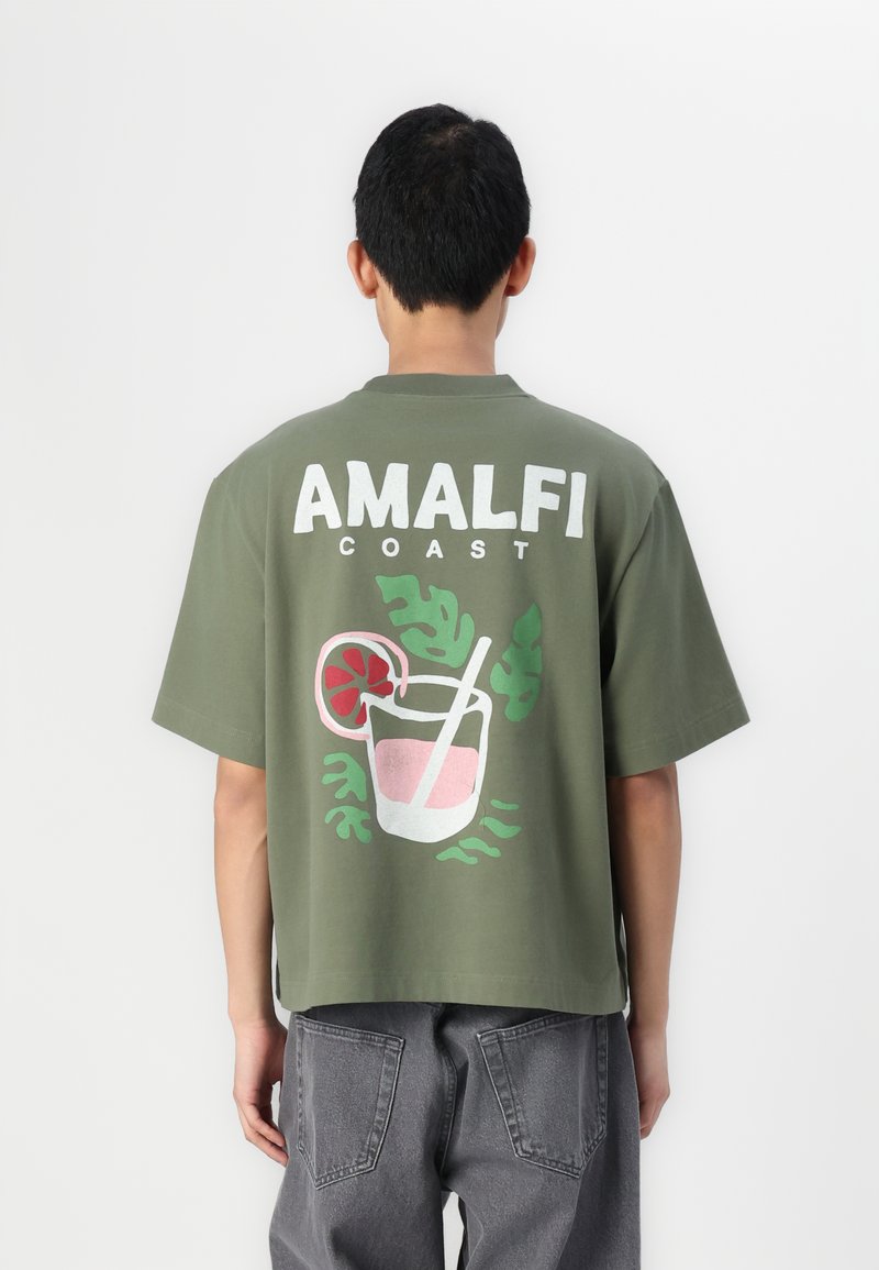 Camiseta de manga corta color verde oliva con el texto en blanco "AMALFI COAST" y un gráfico de un cóctel con hojas en tonos rosa y verde.