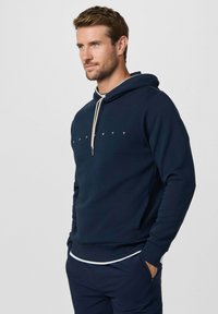 Muž s krátkymi hnedými vlasmi, oblečený v tmavomodrej mikine s bielymi šnúrkami a vhodných nohaviciach, stojaci s rukami v vreckách.