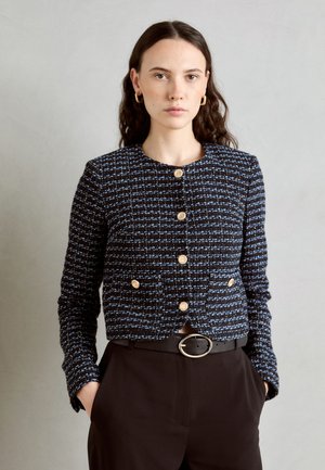 Marineblaues Jacket mit strukturiertem Muster, blauen Akzenten, goldenen Knöpfen und einem cropped Design; kombiniert mit schwarzen Hosen und einem Gürtel.