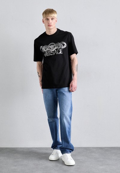T-shirt en coton noir avec un design de logo Versace argenté, associé à un jean en denim bleu clair et des baskets blanches. Modèle debout contre un fond gris.