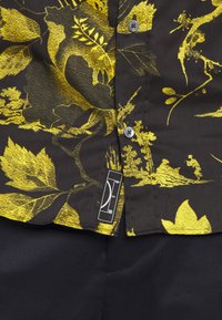 Chemise à motifs floraux noirs et jaunes, boutonnée, avec une petite étiquette rectangulaire noire et blanche près de l'ourlet inférieur, portée sur un pantalon noir.