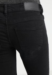 Jeans de mezclilla negra ajustados, con un parche de cuero en la cinturilla y bolsillos traseros estándar con costuras visibles.