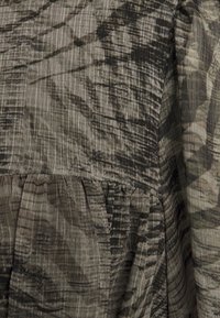 Top in cotone misto caratterizzato da un motivo astratto e texturizzato di colore scuro su uno sfondo verde oliva tenue. Dettagli arricciati all'orlo, sottili righe orizzontali.
