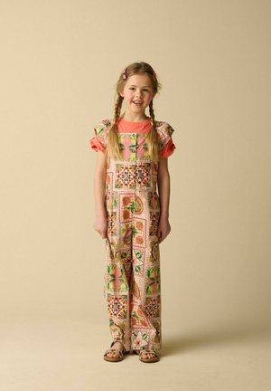 Jeune fille souriante avec des cheveux tressés portant une combinaison à motifs sur un t-shirt corail et des sandales marron, debout sur un fond beige.