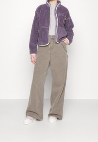 Giacca in pile viola con zip e tasche, abbinata a pantaloni beige a vita alta e gamba larga. Sneakers bianche con dettagli in un viola chiaro.
