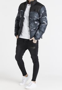 Giacca imbottita con parte superiore nera e parte inferiore in grigio con motivo camo, chiusura con zip, indossata sopra una maglietta bianca. Abbinata a jeans neri e sneakers bianche.