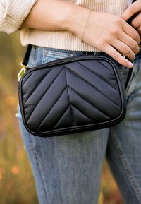 Czarna pikowana skórzana torebka typu crossbody z wzorem chevron, prostokątna forma i złotymi akcentami. Nosi ją przy talii, zestawiona z niebieskimi dżinsami.