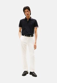 Polo azul marino con cuello contrastante rojo y blanco, combinado con pantalones blancos y zapatos negros. El modelo está de pie con las manos en los bolsillos frente a un fondo neutro.