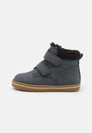 Cizmă cu un exterior din piele albastru închis, două benzi velcro, o margine din fleece negru și o talpă din cauciuc maro, cu cusături albe.