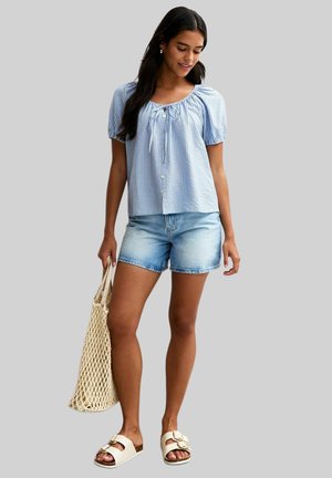 Haut à manches bouffantes rayé bleu avec un décolleté froncé, associé à un short en jean et des sandales à glisser blanches. Sac en filet à la main.