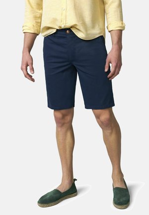 Man draagt marineblauwe korte broek, geel overhemd met lange mouwen opgerold, en groene instappers, staand tegen een witte achtergrond.