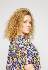 Blusa floral com fundo azul-marinho e flores vibrantes em rosa, amarelo e branco, mangas curtas e um design de corte solto.
