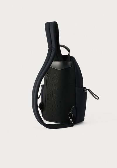 Μαύρο σακίδιο με επενδεδυμένη πλέξη στην πλάτη, ρυθμιζόμενους ιμάντες ώμου, πλαϊνές τσέπες με κορδόνια και επάνω λαβή σε λευκό φόντο.