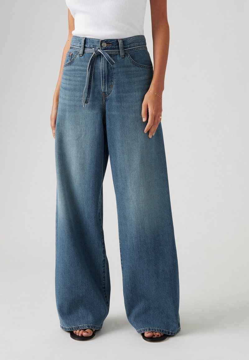 Femme portant un jean bleu taille haute à jambes larges avec ceinture nouée, associé à un débardeur blanc sans manches et des sandales noires à bouts ouverts.