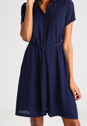 Frau trägt ein knielanges, marineblaues Kleid mit kurzen Ärmeln, einer durchgehenden Knopfleiste und einem gebundenen Taillengürtel.