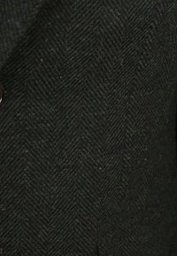 Donkergroen herringbone wol stof met een gestructureerde afwerking. Beschikt over een bruine knoopdetail en zichtbaar stiksel.