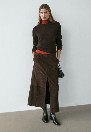 Frau trägt einen dunkelbraunen Pullover über einem roten Rollkragenpullover, einen passenden braunen Midirock mit Frontschlitz, schwarze Stiefel und hält eine schwarze Handtasche.