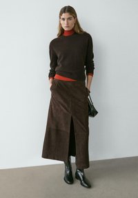 Frau trägt einen dunkelbraunen Pullover über einem roten Rollkragenpullover, einen passenden braunen Midirock mit Frontschlitz, schwarze Stiefel und hält eine schwarze Handtasche.