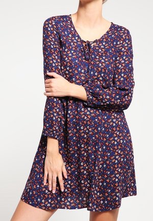 Mujer que lleva un vestido azul marino de manga larga con un pequeño estampado floral y un detalle de lazo en el escote, de pie frente a un fondo liso.