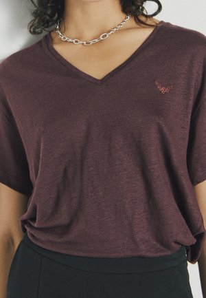 Camiseta de cuello en V color burdeos con mangas cortas, textura suave y un sutil bordado de alas en el lado izquierdo del pecho. Combinada con pantalones negros.