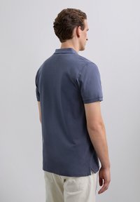 Hombre con una camiseta polo azul de manga corta y pantalones beige claros, de pie ligeramente de espaldas contra un fondo liso.