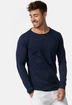 Ung mand iført en marineblå langærmet skjorte og hvide bukser, smilende og kigger ned med den ene hånd i lommen.