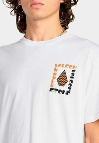Joven con una camiseta blanca que tiene un logo de Volcom Stone en cuadros naranjas y negros en el lado izquierdo del pecho.