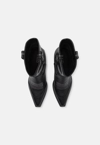 Par črnih usnjenih kavbojskih škornjev z belim šivom in ostrimi konicami, pogled iz zgoraj na beli površini.