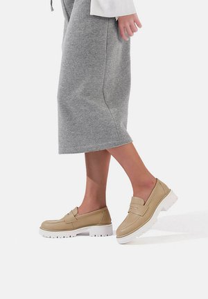 Mocassini beige con una finitura liscia, una banda cucita rialzata e una suola bianca spessa, esposti accanto a una gonna grigia al ginocchio.
