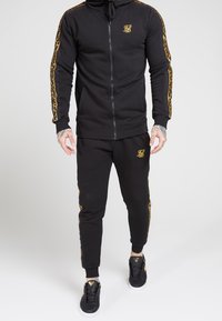 Tuta sportiva nera con dettagli dorati, felpa con cappuccio con zip e logo, pantaloni coordinati e strisce laterali ornate. Indossata con scarpe da ginnastica nere.
